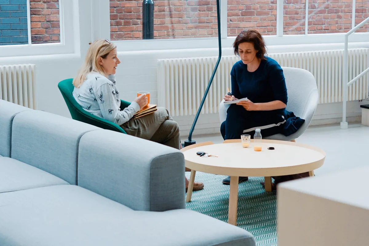 Deux femmes assises face à face dans des fauteuils clairs, l’une prend des notes pendant que l’autre parle
