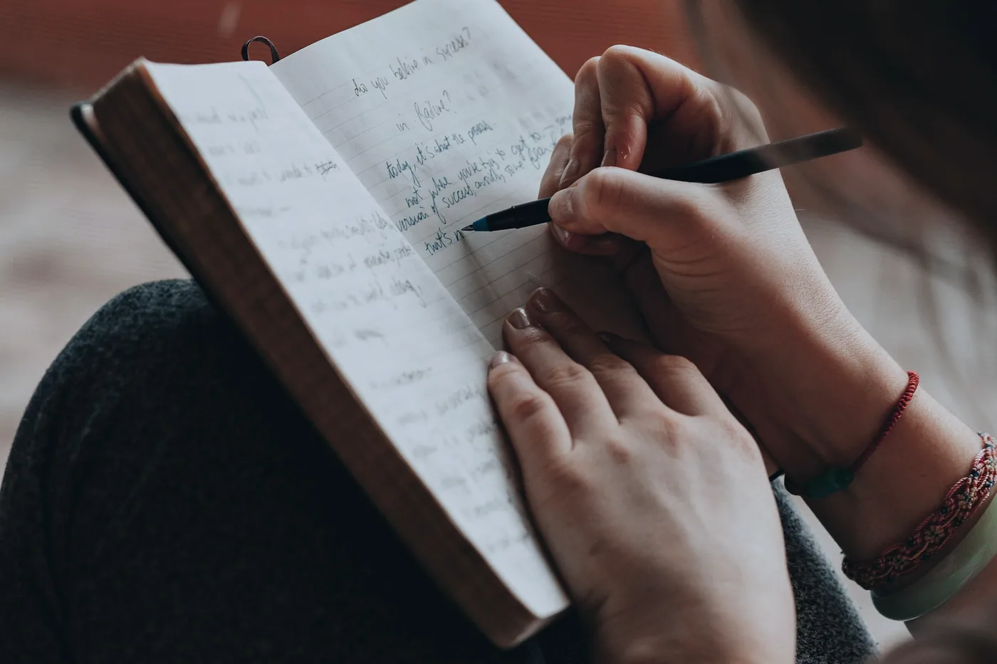 Deux mains écrivent dans un carnet ouvert posé sur les genoux d’une personne vue de dos, un stylo noir à la main