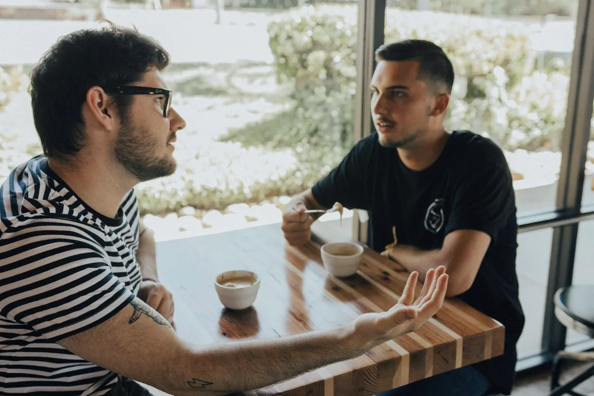 Deux hommes assis face à face à une table de café, l’un accompagne ses paroles d’un geste de la main